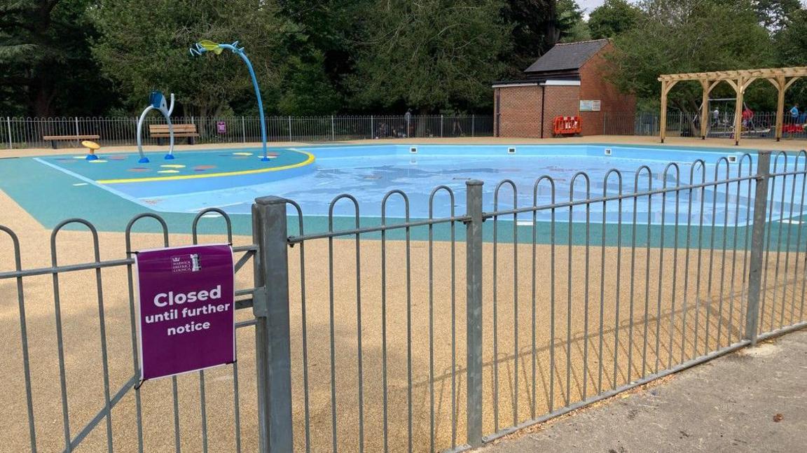 A fenced-off children's paddling pool in a park, with splash pad including mini jets. It is surrounded by green and yellow flooring, with benches around the outside, and trees can be seen beyond. A purple sign on the gate says closed until further notice.