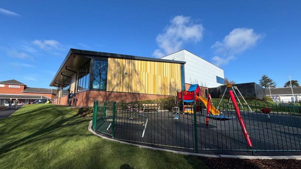 The exterior of a wood-panelled building on a grassy hill with a multi-coloured children's play area in front of it