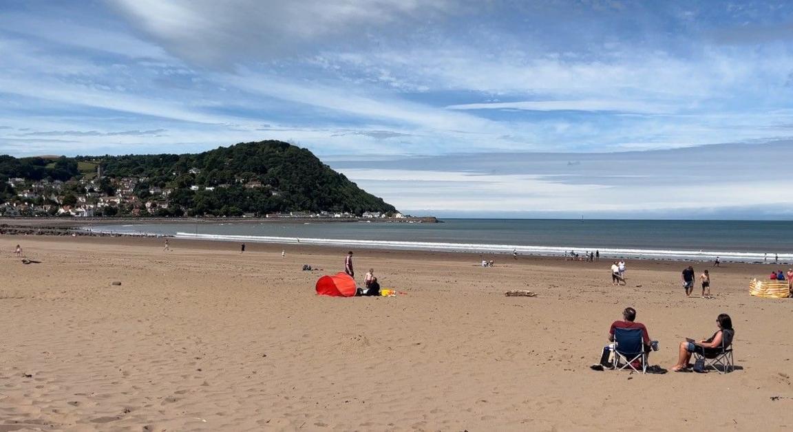 The beach at Minehead with a few people sat or walking on the sand. The tide is in and you can see North Hill in the distance.