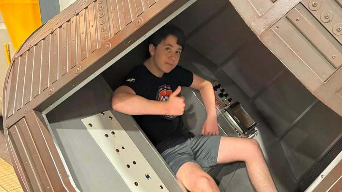 Aston lying down in a space shuttle. He has dark hair and is wearing a dark T-shirt and grey shorts. He is giving the thumbs up.
