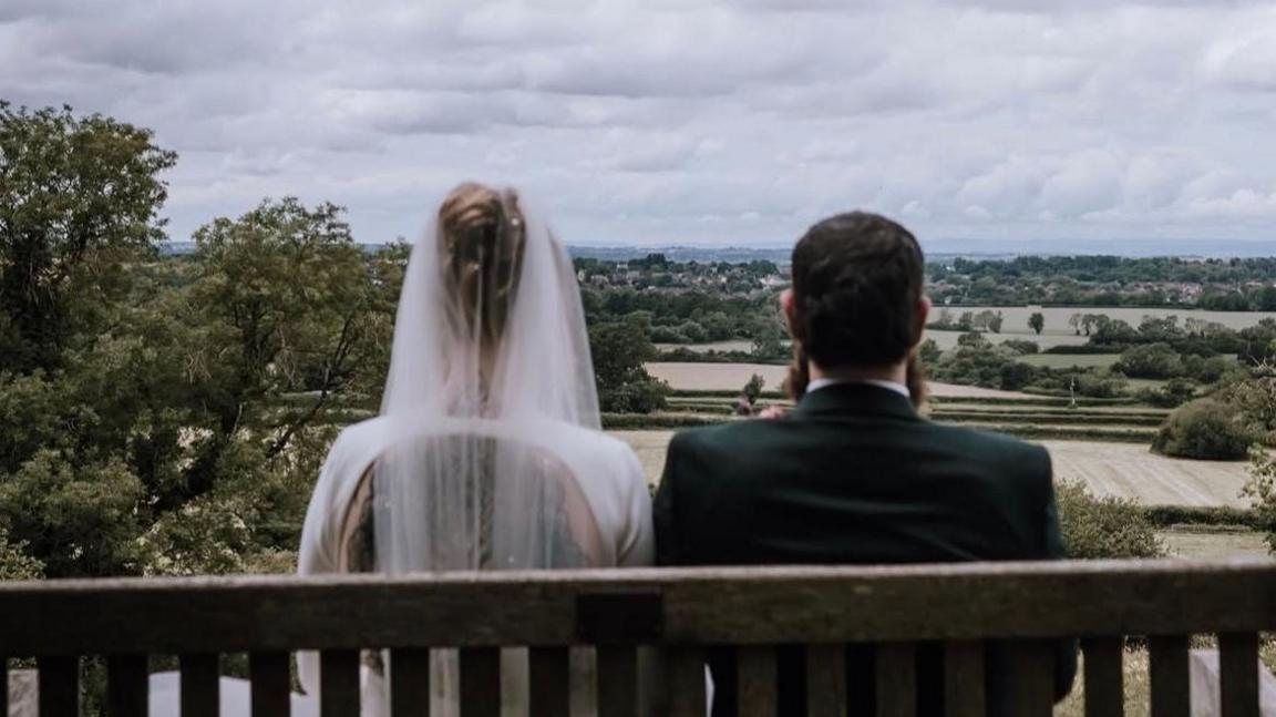 A bride and groom sit side-by-side on a wooden bench with their backs to the camera, looking at the view of fields. The groom is wearing a dark suit. The bride is wearing a white dress.