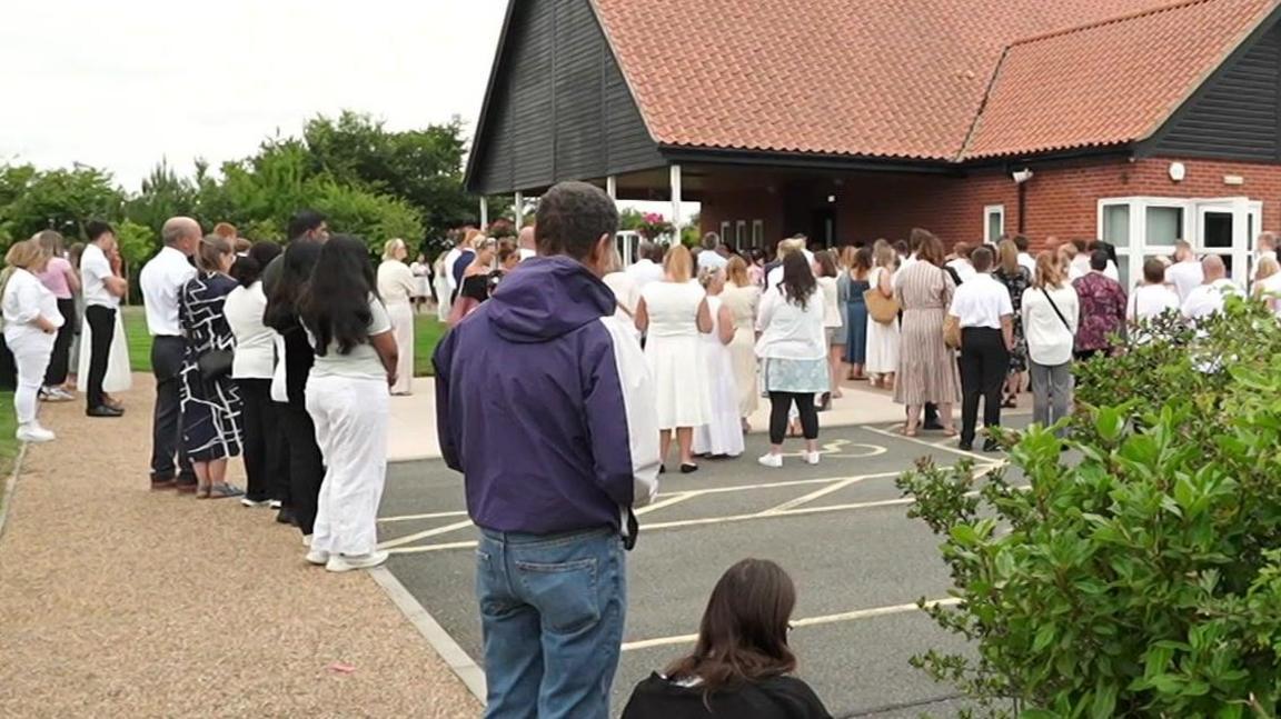 Lots of people are dressed in white standing on parts of a car park and walk way outside of a stone coloured building, a crematorium, which is where the funeral was taking place.