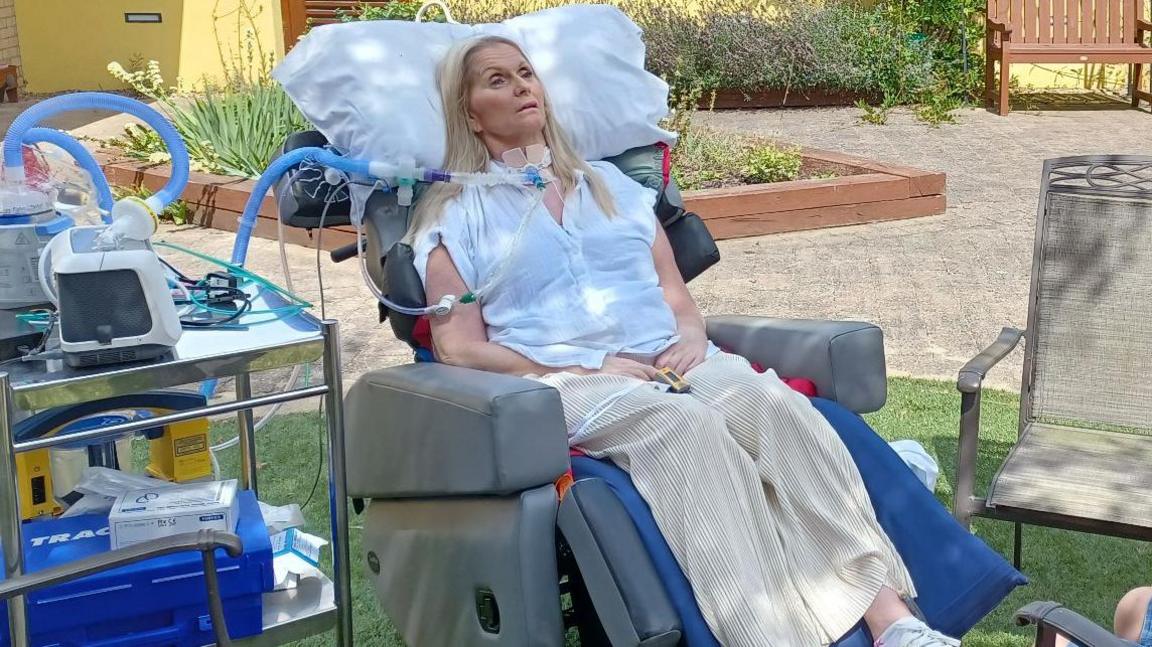 Catherine lying in a medical bed in a garden. She is connected to medical equipment. A young boy is sat by the bed.