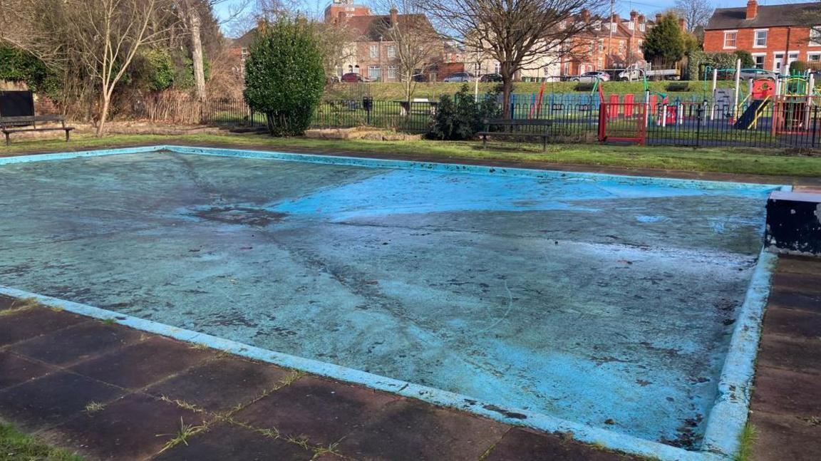 The closed-off paddling pool in St George's park lies empty on a sunny day. Dirt and moss are visible on the blue paint of the pool's floor.