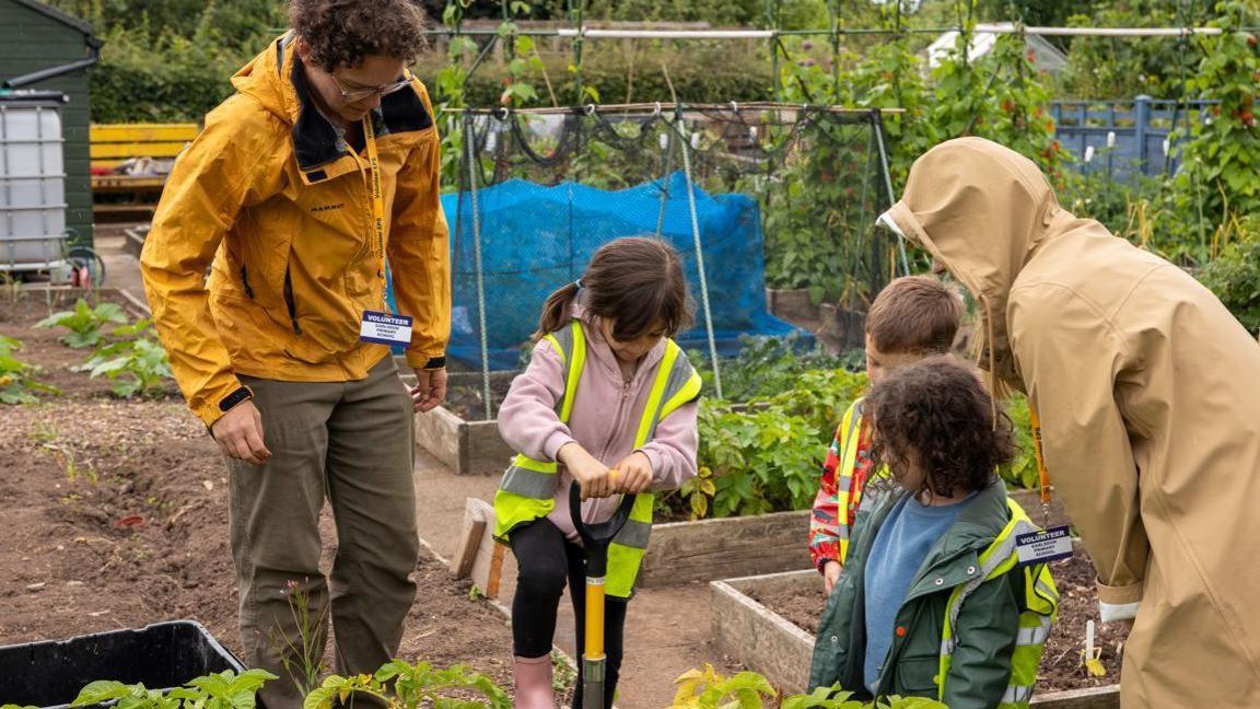 Three children at the plot. All are wearing hi-visibility green jackets and a girl is using a spade. A man and a woman are supervising and plants can be seen growing. 