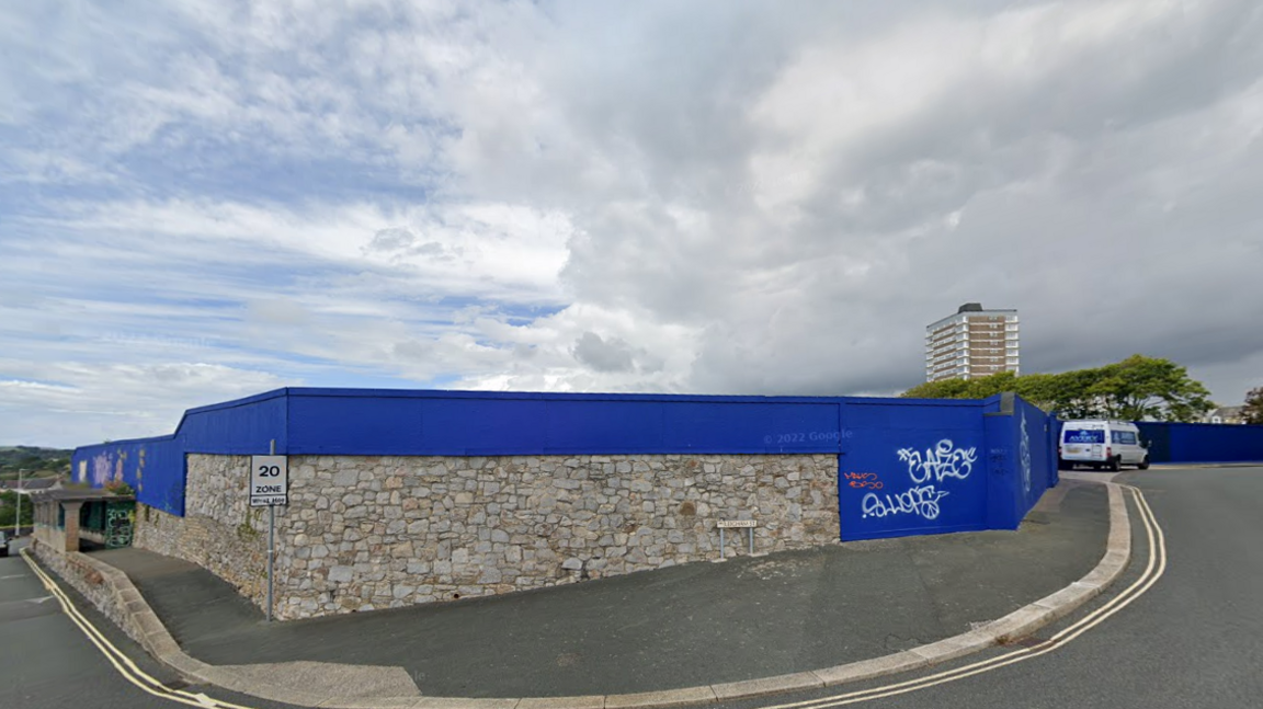 A Google street view screen grab of the derelict hotel site, hidden by tall navy panels, cloudy skies