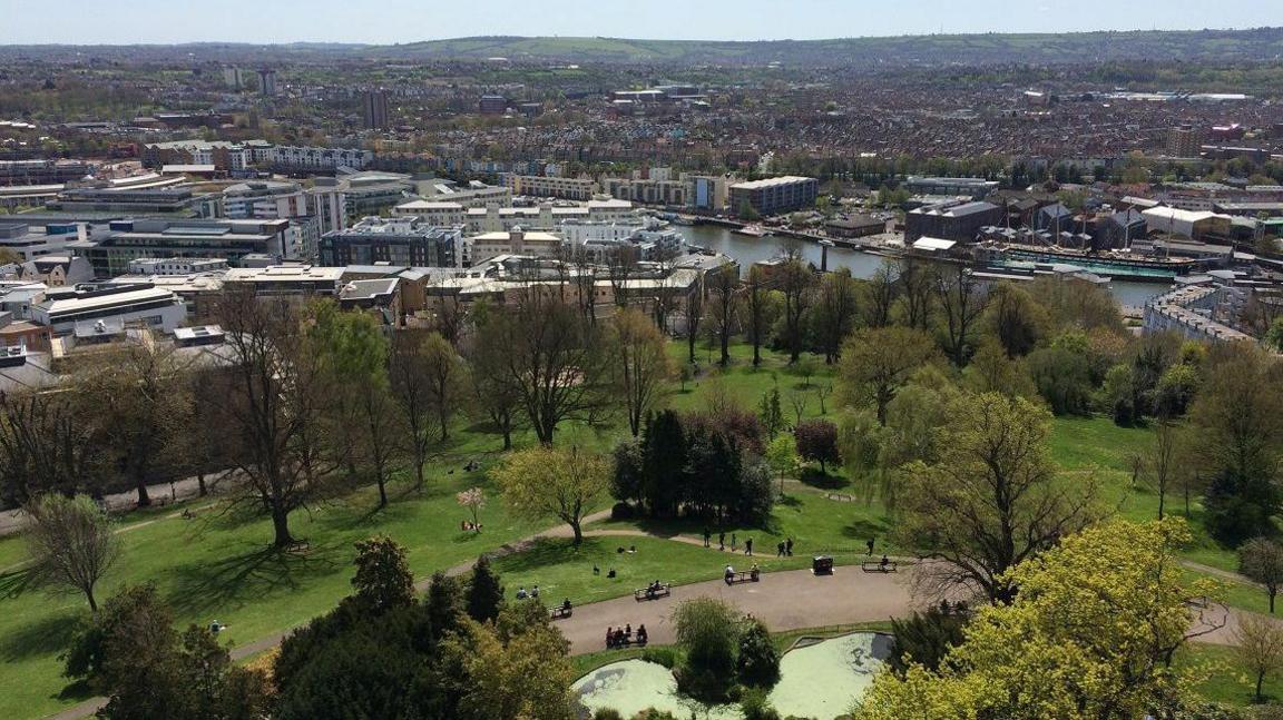 Aerial view of a cityscape with a large park in the foreground, featuring green lawns, trees, walking paths, and people relaxing. A river with docked boats separates the park from a mix of residential and commercial buildings. Rolling hills are visible in the distance under a clear sky