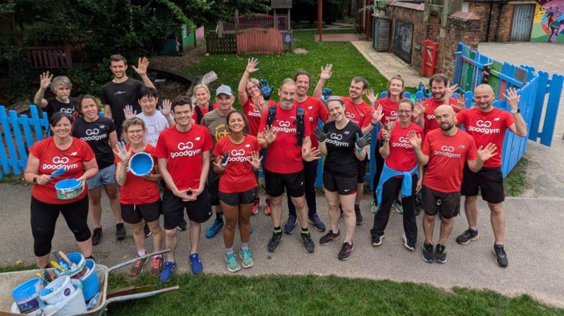 About 18 people stand in a school garden painting fences blue. They are wearing red and black GoodGym T shirts, shorts and trainers.