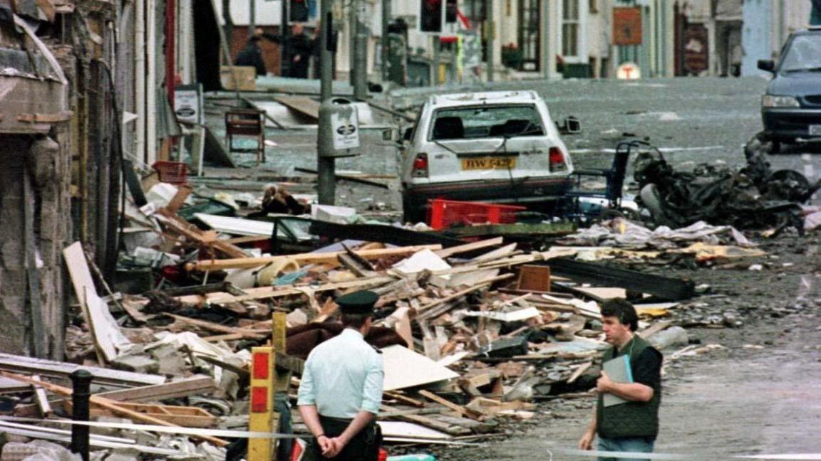 Archive photo of the aftermath of the Omagh bomb in August 1998.  Piles of wood and rubble lie on the street outside damaged shops.  The wreckage of a car and a damaged wheelchair are in the background.  A uniformed RUC office stands at a cord with his hands behind his back.  Another man wearing a headset and holding a book is talking to the officer. 