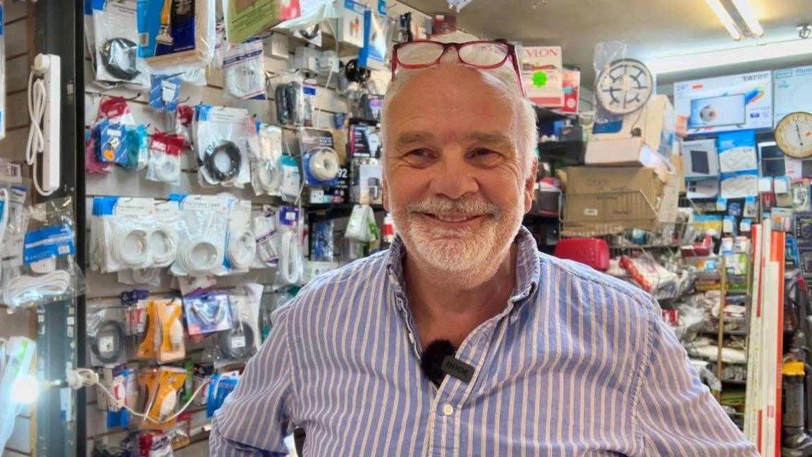 A man in a striped top looks into the camera. He has short white hair with his red-rimmed glasses perched on his forehead. He has a short white goatee beard. Behind him, a range of electrical and hardware items are on pegs in his market stall.