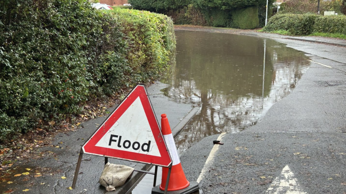 A triangular flood warning sign is positioned in front of a large flooded section of road.