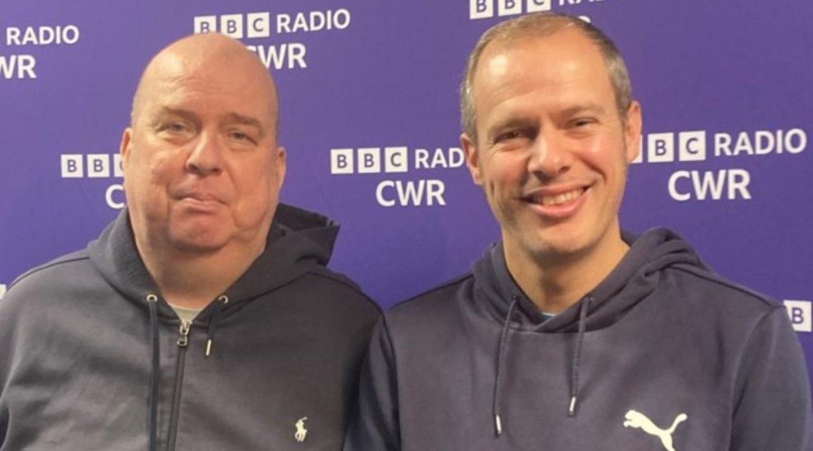 Neil Jeffs on the left is standing next to younger brother Paul, who is smiling at the camera. They are in front of BBC Radio CWR branding