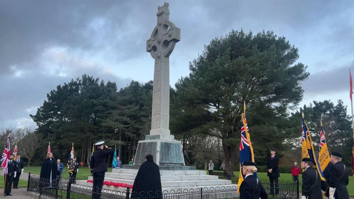 A man salutes at the foot of the national memorial in St John's. It is a tall sculpture in the shape of Celtic cross with Celtic designs on all four sides. Standard bearers are holding standards for the armed forces and other organisations on either side.