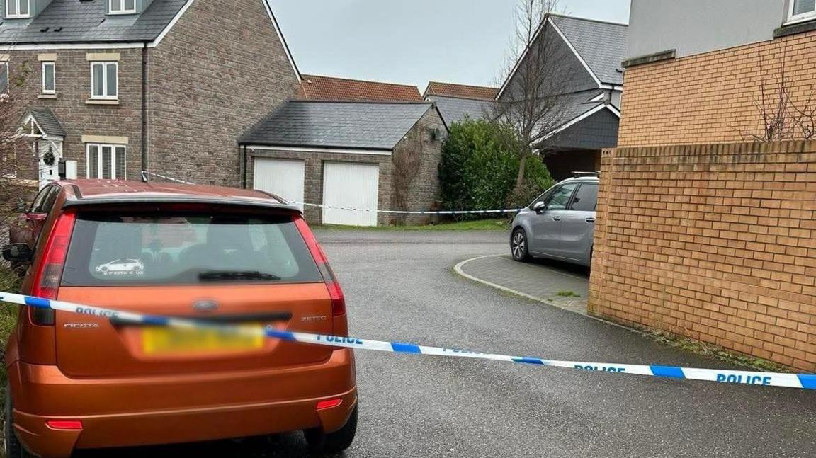 Police cordon tape is stretched across two section of a road in a residential estate. There are two parked cars in sight and a large house with a double garage.