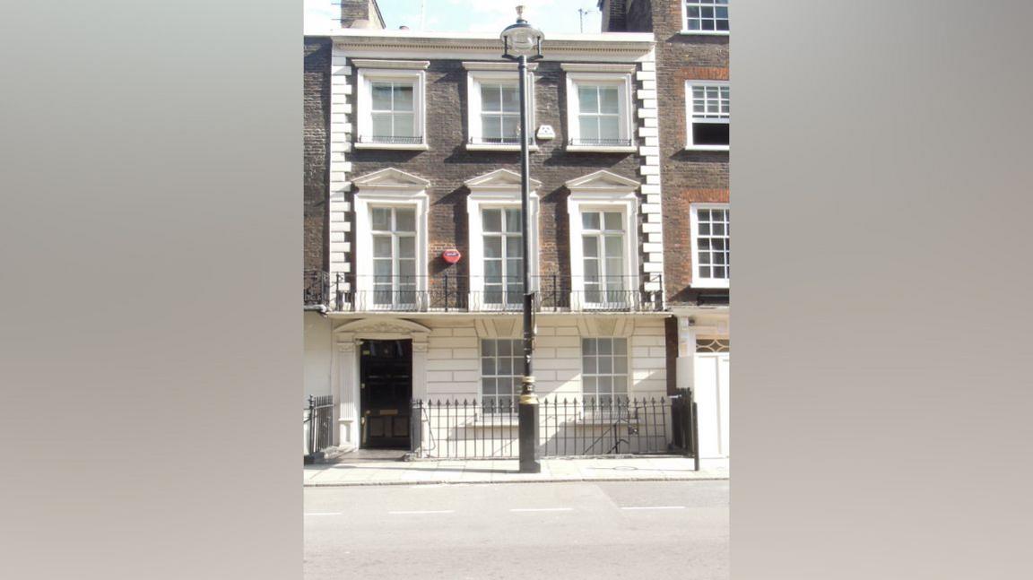Front view of a 18th Century brick townhouse in London. It has a black front door and appears to be three storeys tall. There is a traditional black lamppost on the pavement outside