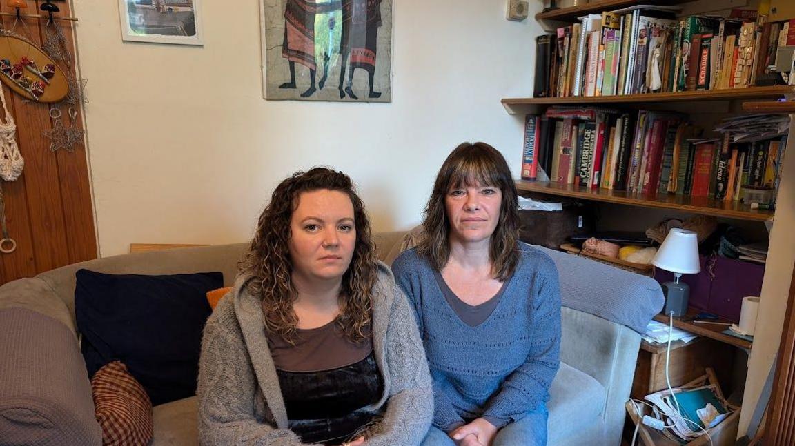 A picture of Juliet Rimer with long, curly, brown hair and wearing a black top and grey cardigan. She is sat on a sofa next to a bookshelf alongside Kate Rimer, who has long, brown hair and is wearing jeans and a blue jumper.