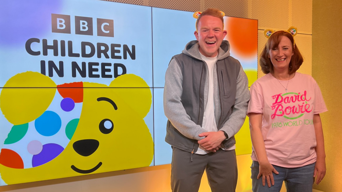 Colson Smith and Gayle Lofthouse indoors in front of a large digital display. The display features the BBC Children in Need logo, which includes the text “BBC Children in Need” and an illustration of Pudsey Bear. The background of the display is bright and well-lit.
Colson on the left is wearing a light grey jacket over a white hoodie, paired with grey trousers. Gayle is wearing a pink T-shirt with the words “David Bowie” printed on it in bright colours, along with blue jeans.