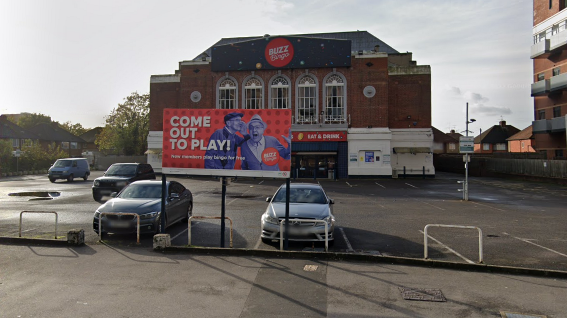 A general view photo of a bingo hall, with Buzz Bingo branding at the top of it, and four cars parked in a car park. The building looks like a fairly grand early 20th century building with five tall windows at the front of it 