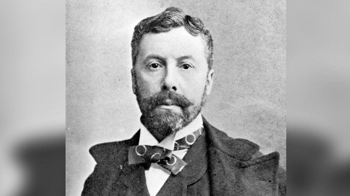 A black and white photo of Richard D'Oyly Carte who has a beard and moustache and is wearing a heavy suit with a bow tie. He is looking at the camera and his dark hair has a side parting.