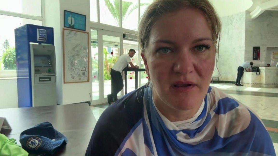 A woman has taken a selfie in an airport. Behind her are a few security staff and safety barriers. There is also an ATM in the background. 