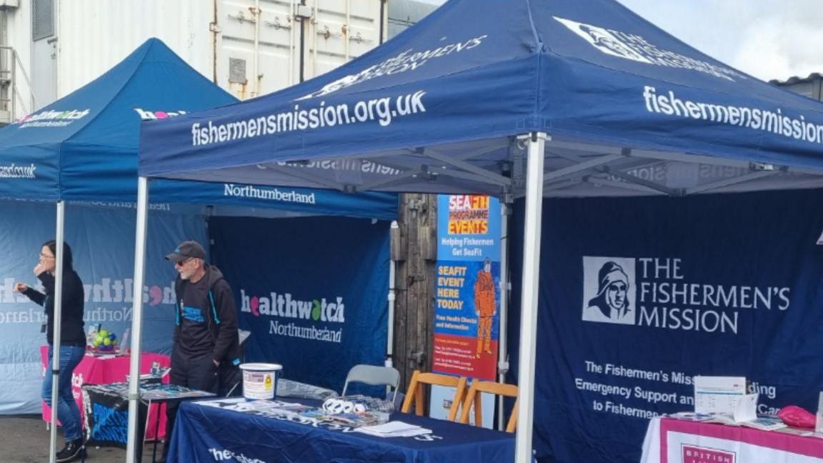 Blue gazebos with branding saying: 'SeaFit', 'The Fishermen's mission' and 'Healthwatch Northumberland'. There are tables with leaflets and medical equipment and two people manning the stalls.