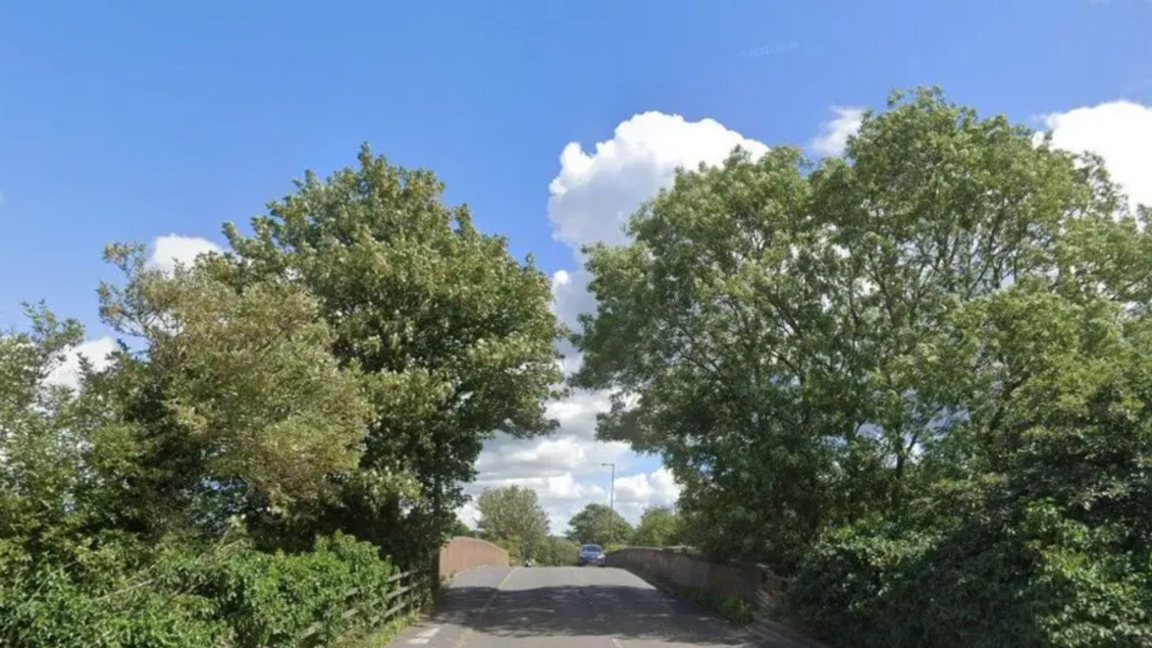 A road, with trees overhanging from either side, and a pavement on the left hand side.
