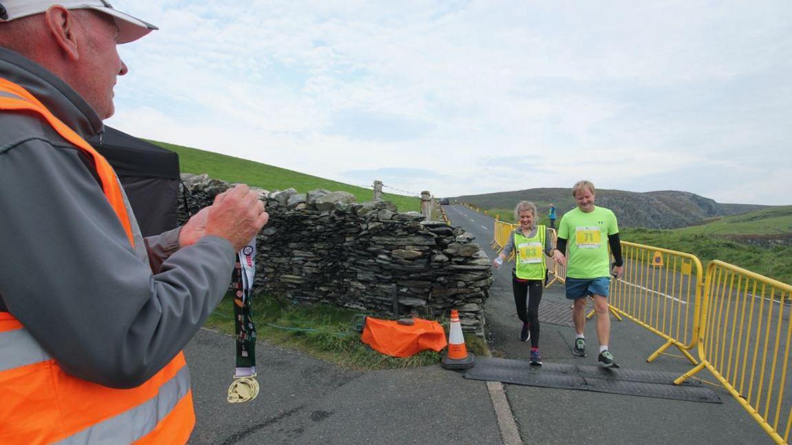 Two walkers hold hands while crossing the finish line at the Sound. They are walking across a black mat next to yellow barriers while a marshal in an orange hi-vis waistcoat holds out medals for them.