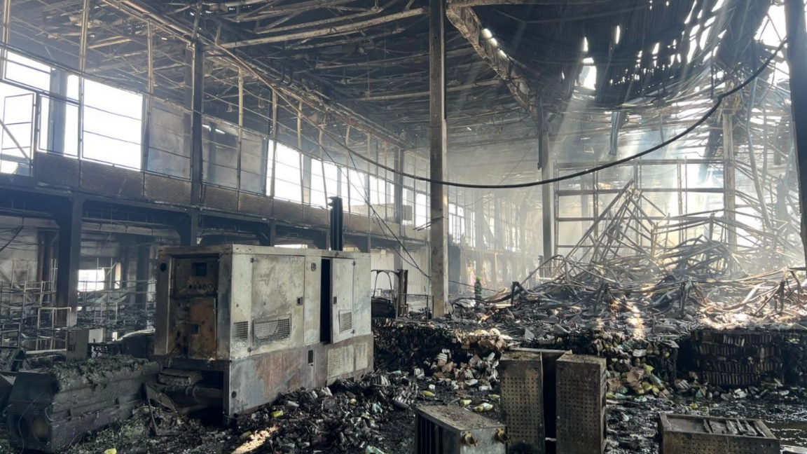 The warehouse as seen from the inside - light pours in from holes in the ceiling and walls, and there is charred debris spread across the ground.