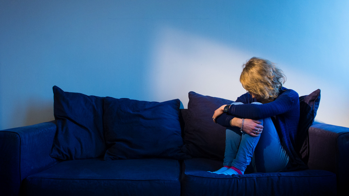 A woman huddled up in the corner of a sofa with her hands across her knees and her head down