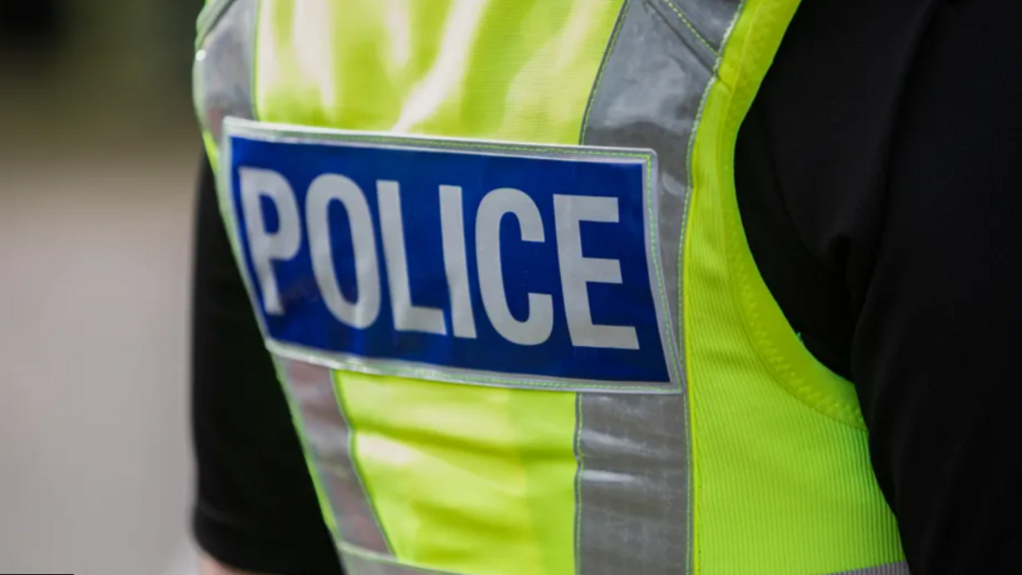 The back view of a British police officer's yellow vest. It says "POLICE" in white block lettering against a blue rectangular background  