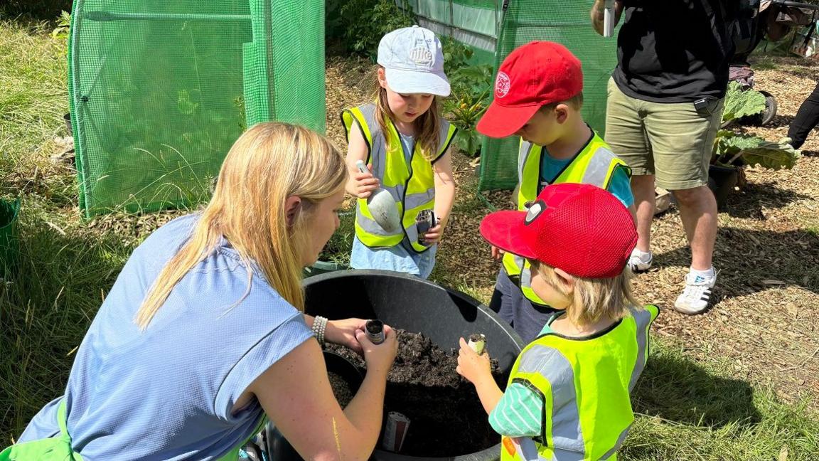 A woman with long blonde hair wearing a blue T-shirt is kneeling next to a bucket of soil with three children in yellow hi-vis vests. Two are wearing red caps and one has a white cap. There is a green growing tunnel behind them.