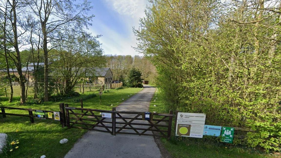 The entrance to the donkey sanctuary site. It shows a single road leading into the distance with grass and trees on either side. There is a closed wooden gate on the road and the fencing to the right has three signs, one labelled 'The Donkey Sanctuary'. There is a barn in the distance on the left. The sky is blue with light clouds. 