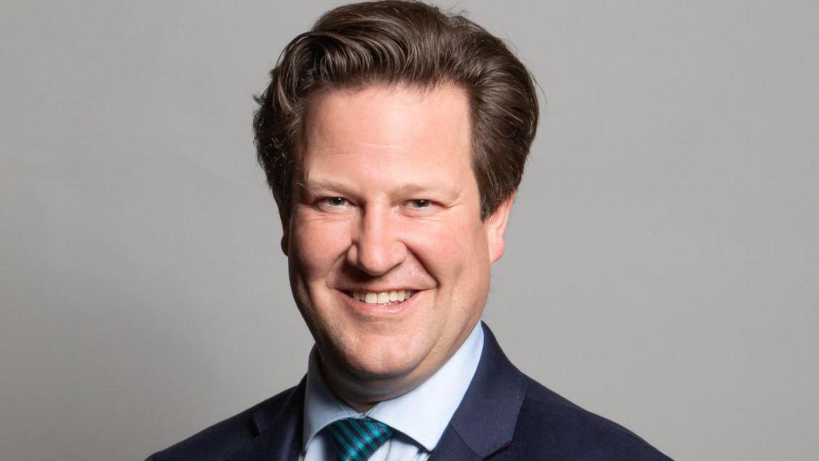 A man in a navy blue suit and a pale blue shirt with a green tie, smiling against a grey backdrop.
