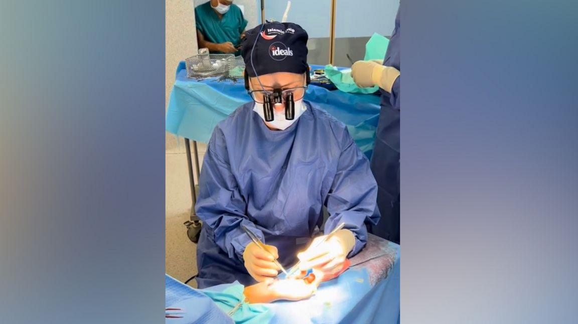 Victoria wears operating scrubs and goggles whilst operating on a hand under a spotlight 