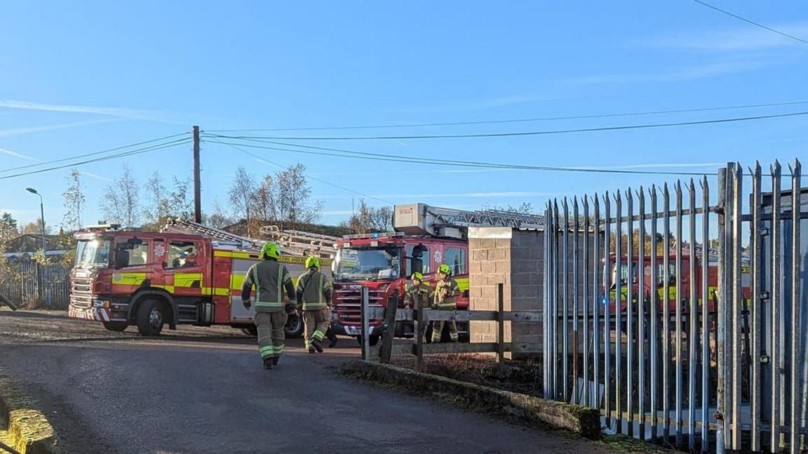 Three fire engines are parked on a road by an industrial estate. Four firefighters can be seen walking in front of the fire engines. The area is otherwise quiet and the sky is a clear blue and bright.