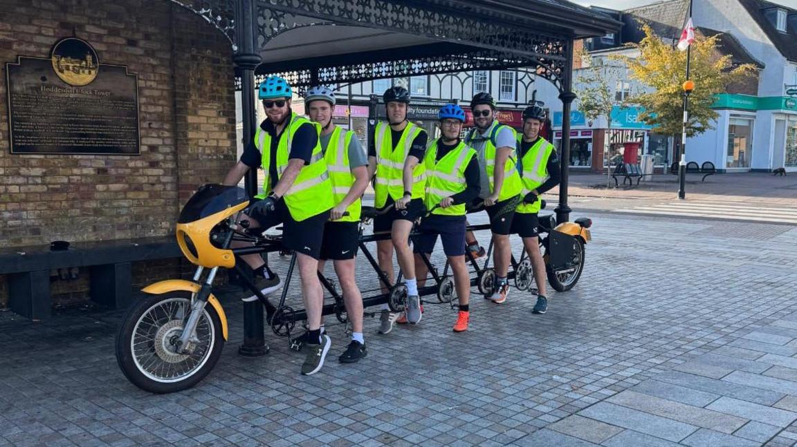 Six men on a very long bicycle which has six seats and six sets of pedals. They are wearing yellow high vis jackets