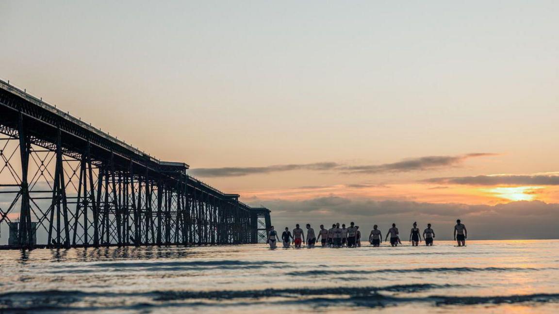 A group of swimmers in trunks and swimming costumes walking out of the sea at Ramsey at sunrise, with the water level is up to their knees. The Queen's Pier, which is a long iron pier, can be seen in the background in silhouette. The sky has pink hues.