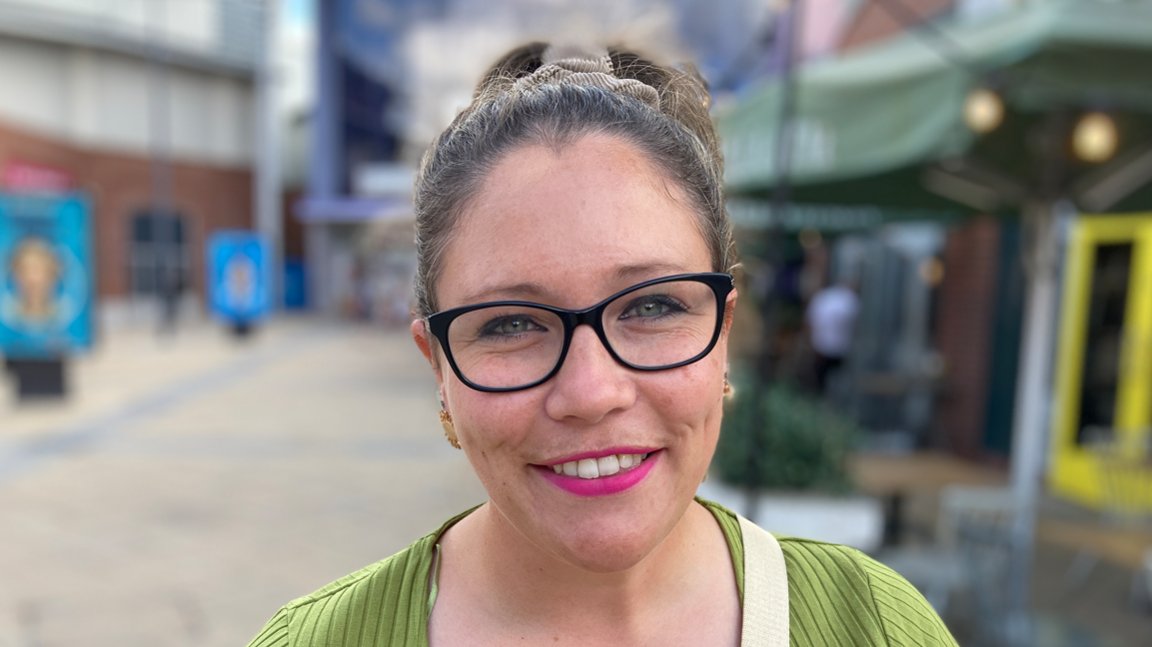 A head and shoulders image of Sarah Catton. She is wearing a green jumper and glasses. She is smiling into the camera.