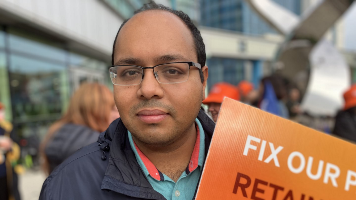 A man wearing glasses holds an orange sign that is mostly out of shot, the words "fix our" are visible. The background is out of focus but it is clear that he is standing with a group of people in front of a set of buildings