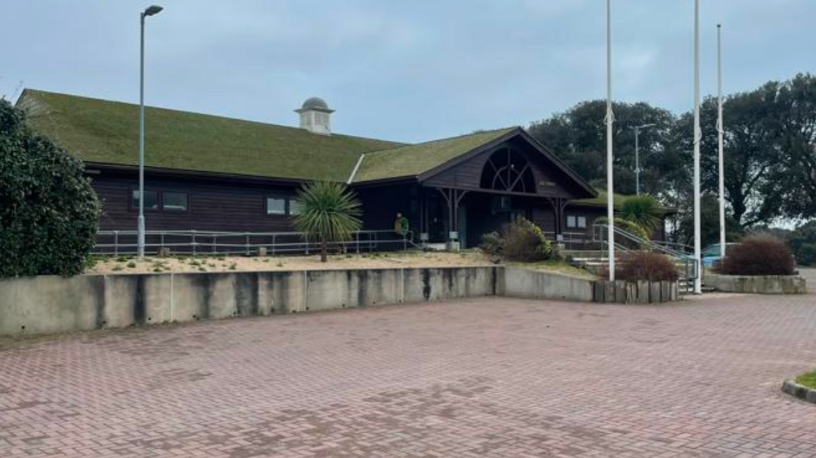 A building with a large roof and dark frontage and a paved area in front along with a number of flagpoles.