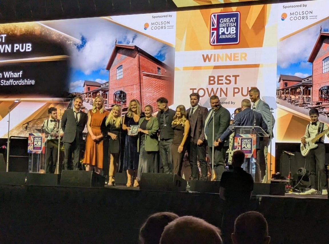 Fourteen people, men and women, are on stage. Some of the women are holding an award. The words Winner Best Town Pub are on the large vertical display behind them.