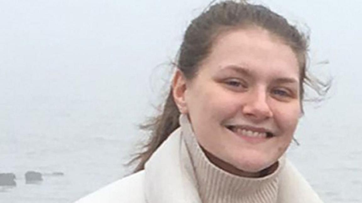 Libby Squire smiling for the camera. She appears to be at the seaside on a cold and cloudy day.