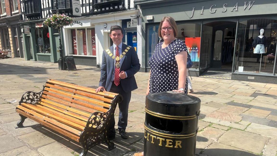 A man with dark hair in a blue suit and red tie and mayoral chain standing behind a wooden slatted bench with black metal sides. Next to him, a woman with brown hair and a black dress with white spots is standing behind a black metal litter bin with the word litter in gold writing. They are on a paved street with shop fronts in the background.