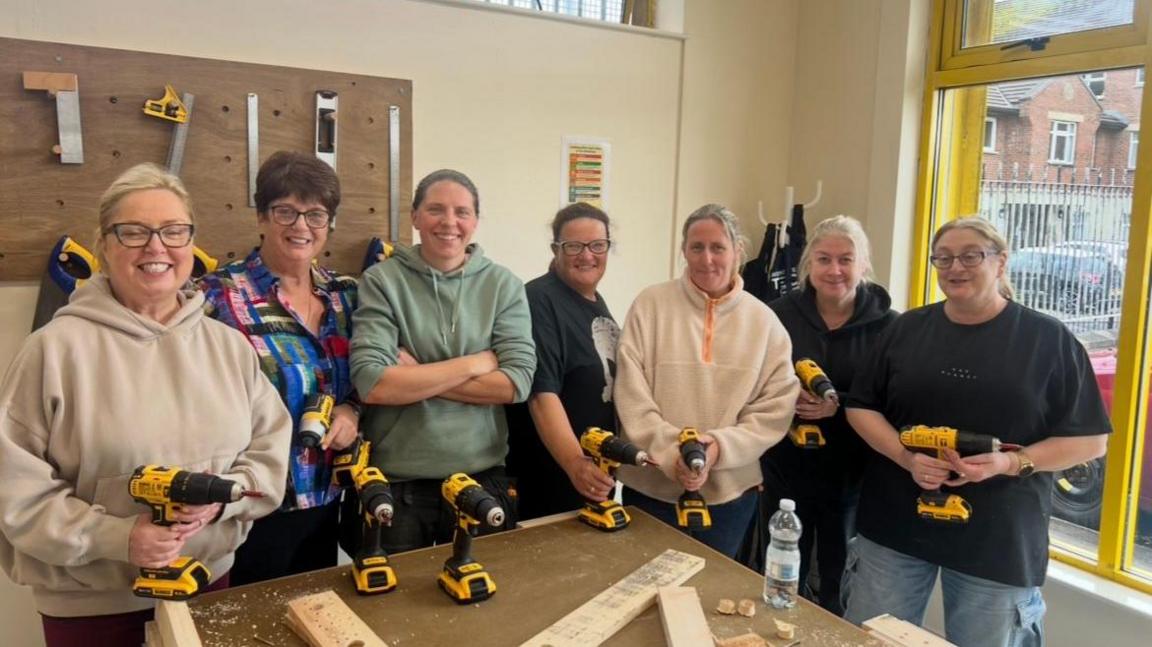 Women standing in different coloured jumpers holding electrical drills. They are standing around a table that has pieces of wood on it. 