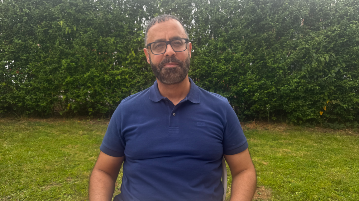Mohammed Bashir is sat down on a chair in a garden with a neutral facial expression. He is wearing wide black frame glasses and a blue open collar t-shirt. He has short black hair and a black beard 