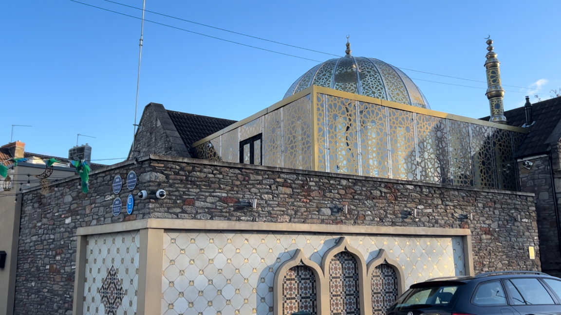 An exterior view of Easton Jamia Masjid. The building has an exterior brick wall and the internal section of the mosque has patterned windows and an ornate dome.