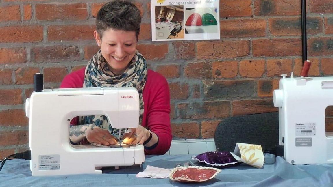 A smiling woman sitting working on a sewing machine. She wears a pink top and a cravat style scarf. She has short, dark pixie style hair.