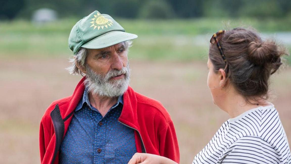 A photo of Angus Wainwright on the site of the dig at Sutton Hoo. He is in a field speaking with a women. He is facing her while in mid-conversation. He has a grey beard and is wearing a green cap. He is also wearing a red jacket with a blue shirt underneath.