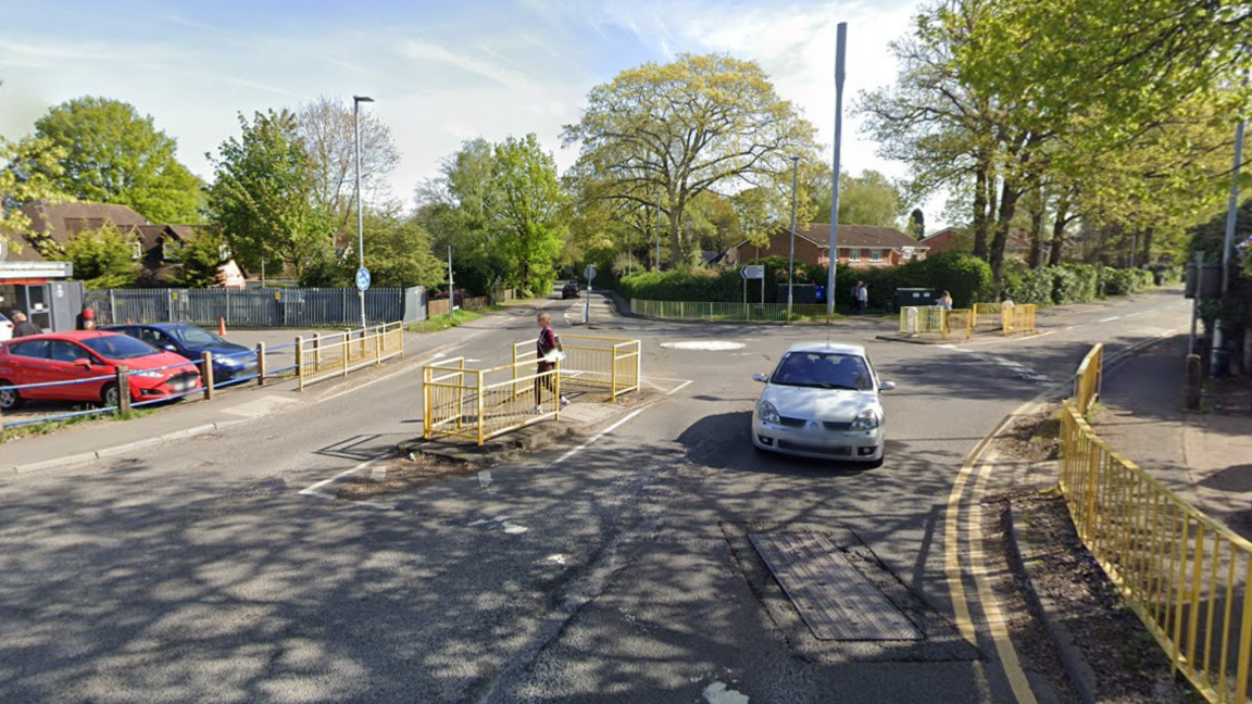 A screenshot from Google street view showing what the area used to look like. There's a mini roundabout with a white painted island in the middle. There are no crossings at the exits, just gaps in the yellow pavement railings suggesting people can cross there.