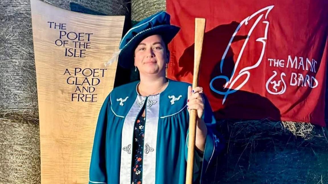 Jordanne Kennaugh wearing a blue gown with a flamboyant traditional hat of a bard and holding a pole, in front of a sign that reads the poet of the isle, a poet glad and free.